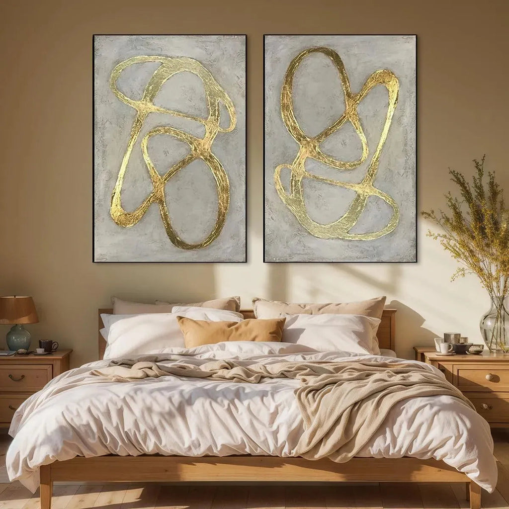 Two vertical abstract paintings with textured gray backgrounds and bold raised gold lines, displayed above a wooden bed.